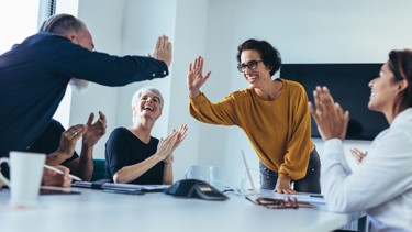 Five colleagues celebrating around a meeting table with high-fives and clapping in a modern office