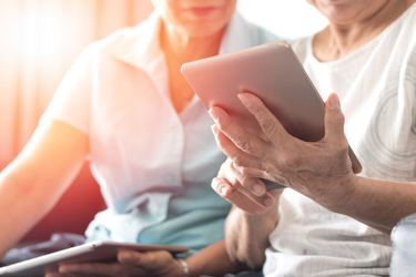 an elderly person and a younger lady sitting on a couch while looking at the screen of a tablet