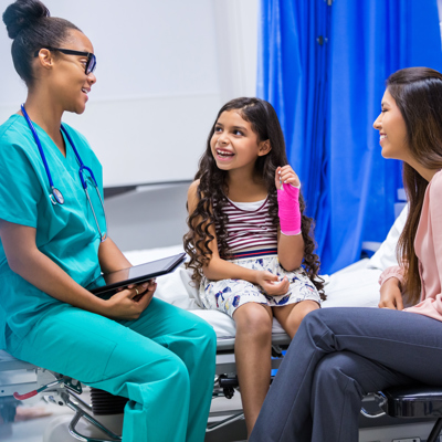 Nurse sitting on the bed with a young child who's arm is broken and her mother sitting down on a chair besides her smiling