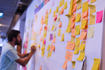 A large whiteboard filled with post-it notes while a man is writing a new one