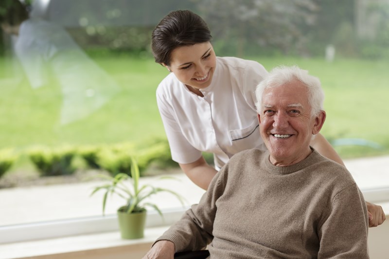 An elderly man smiling at the camera sitting down while a female carer has her hand on his shoulder and smiles at him