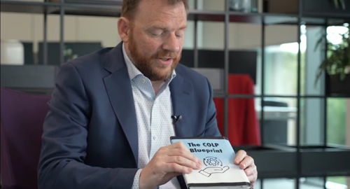 Man in a blue suit holding and discussing a book titled 'The COLP Blueprint.'